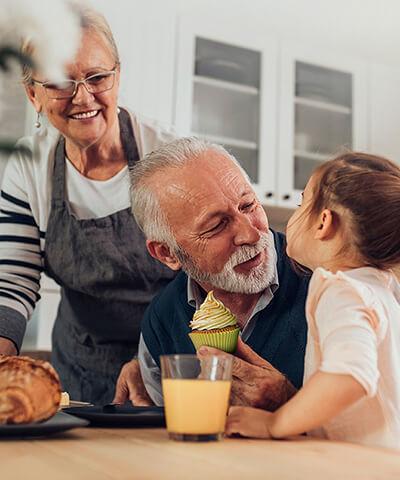 Porque somos asi Pensiones - Pareja senior con su nieta pequeña cocinando magdalenas y sonriendo en la cocina de su casa