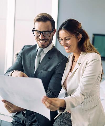 Momento Hacer crecer mi empresa - Hombre y mujer de negocios con traje, corbata y gafas sonriendo mientras miran papeles en la oficina